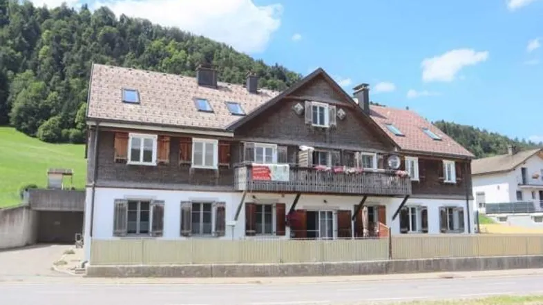 Mehrstöckiges Gebäude mit traditioneller Holzfassade, Balkon und Ziegeldach vor grüner Hügellandschaft.