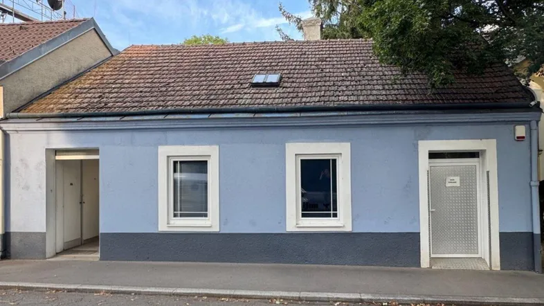 Die Fassade des Hauses zeigt eine hellblaue und dunkelgraue Farbgebung mit zwei Fenstern und einem Eingang.
