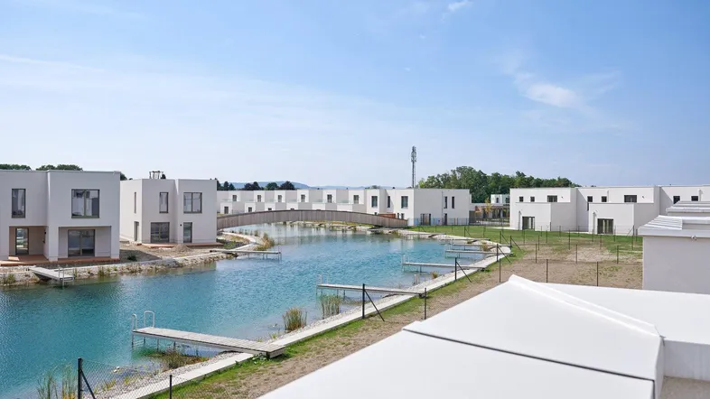 Moderne Reihenhäuser an einer künstlichen Wasserlandschaft mit Brücke unter blauem Himmel.