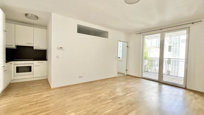 Heller offener Wohnbereich mit integrierter Küchenzeile, Parkettboden und Zugang zum Balkon.