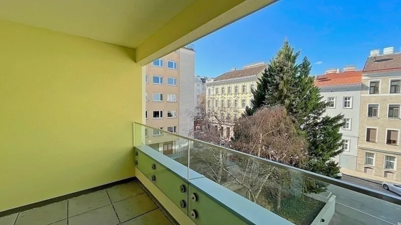 Heller Balkon mit Glasgeländer und Blick auf die umliegenden Gebäude und Bäume.