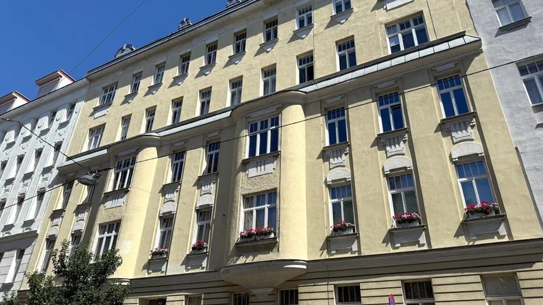 Historische Fassade eines Mehrfamilienhauses mit vielen Fenstern und Blumenkästen unter blauem Himmel.