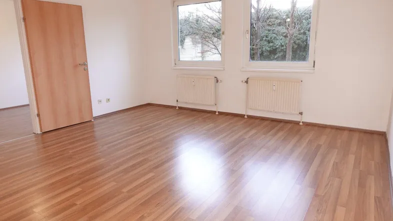 Helles Zimmer mit Laminatboden, weißen Wänden und zwei Fenstern, die viel Tageslicht hereinlassen.
