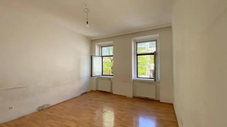 Leeres Zimmer mit Parkettboden, zwei Fenstern und Renovierungsbedarf, ideal zur individuellen Gestaltung.
