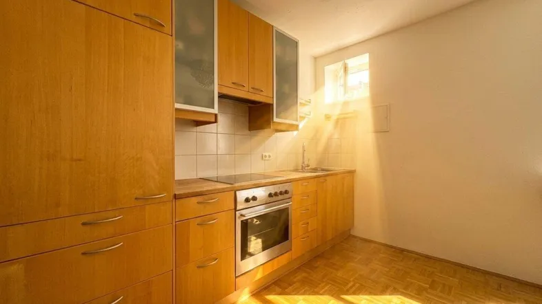 Funktionale Küche mit Holzschränken, Einbaugeräten und hellem Parkettboden, gut beleuchtet durch Sonnenlicht.