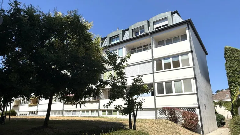 Mehrstöckiges Wohngebäude mit weißer Fassade, Balkonen und grüner Umgebung unter blauem Himmel.