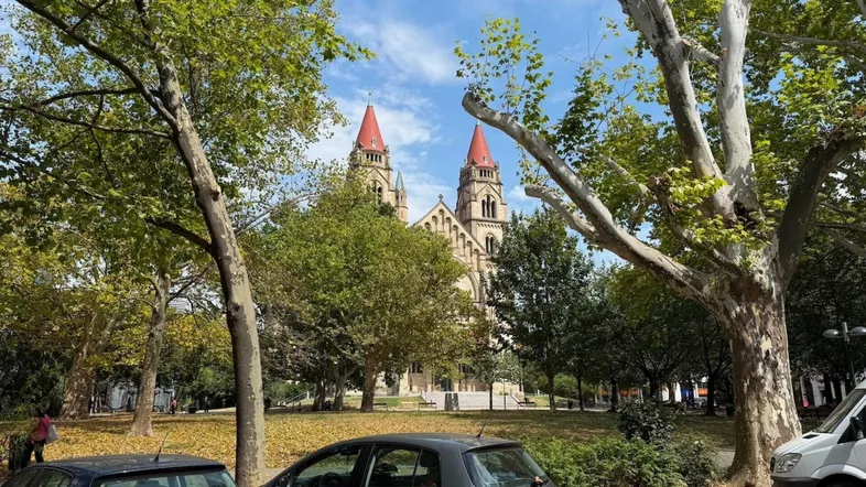 Blick auf eine historische Kirche mit roten Türmen, umgeben von Bäumen und parkenden Autos.