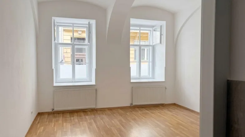 Helles Zimmer mit Holzboden, zwei großen Fenstern und einer eleganten Gewölbedecke.