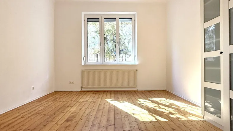 Helles Zimmer mit Holzdielenboden, weißen Wänden und einem großen Fenster, das viel Tageslicht hereinlässt.