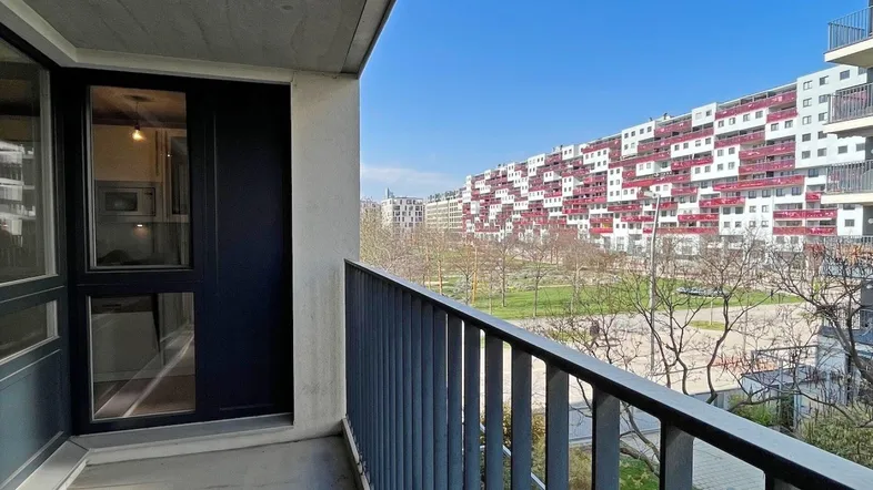 Blick vom Balkon auf moderne Wohngebäude und eine grüne Parkanlage unter blauem Himmel.
