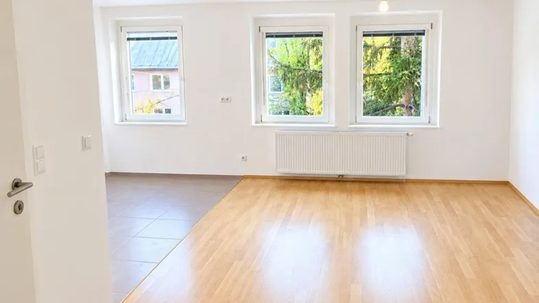 Helles Zimmer mit Parkettboden und Fliesenbereich, ausgestattet mit Heizkörper und Blick ins Grüne.