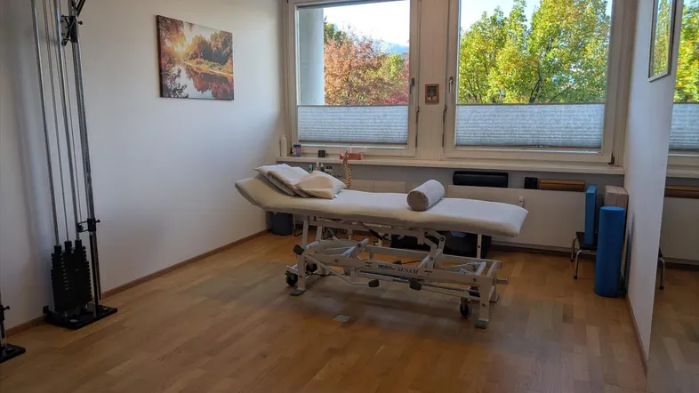 Heller Therapieraum mit Behandlungsliege, Trainingsgerät und großen Fenstern mit Blick ins Grüne.