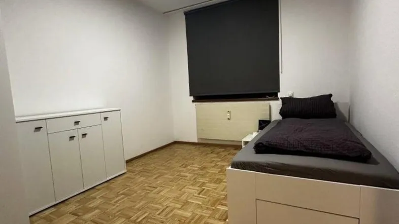 Helles Schlafzimmer mit Parkettboden, einem Bett mit Stauraum und einem Fenster mit Rollladen.