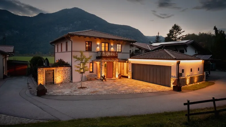 Elegantes Haus bei Dämmerung mit beleuchtetem Eingangsbereich und gepflastertem Vorplatz vor Bergkulisse.