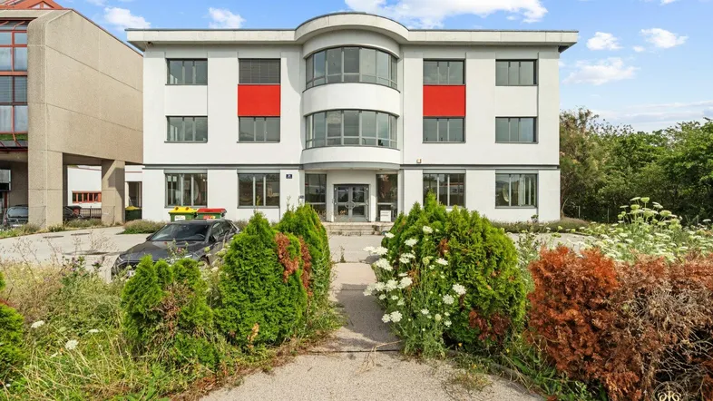 Modernes Bürogebäude mit weißer Fassade, roten Akzenten und gepflegter Grünanlage vor dem Eingang.