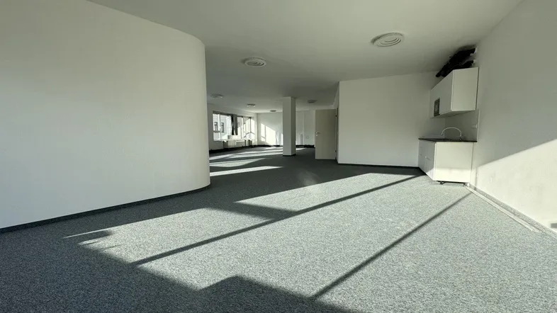 Geräumiges, leeres Büro mit Teppichboden und viel natürlichem Licht durch große Fenster.
