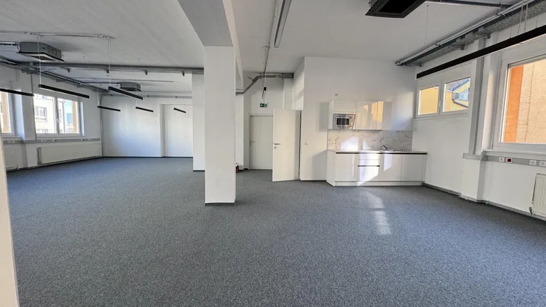 Großer, offener Büroraum mit Teppichboden, weißen Wänden und einer integrierten Küchenzeile, ideal für moderne Arbeitsumgebungen.