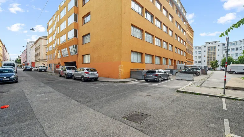 Gebäudeansicht von der Straße mit parkenden Autos und umliegender Stadtarchitektur unter blauem Himmel.