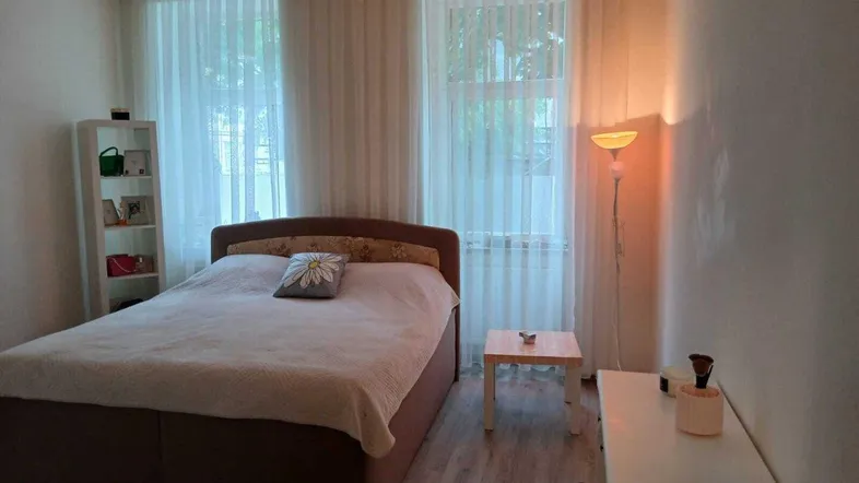 Helles Schlafzimmer mit großem Bett, Laminatboden und zwei hohen Fenstern, die viel Tageslicht hereinlassen.