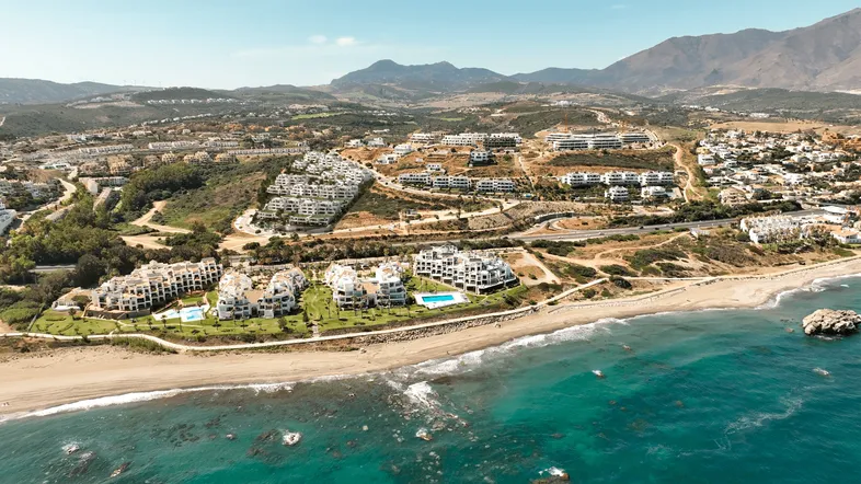 Panoramablick auf eine moderne Wohnanlage am Meer mit Pools, Strand und bergiger Landschaft im Hintergrund.