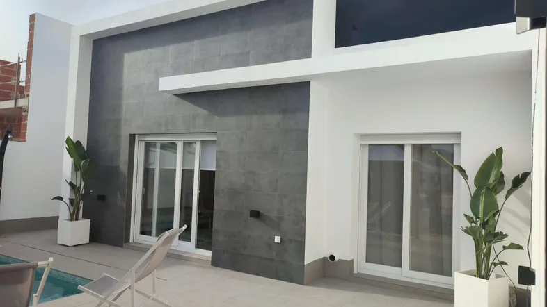 Moderne Hausfassade mit grauem Fliesenakzent, weißer Wand und kleinem Poolbereich mit Liegestühlen.