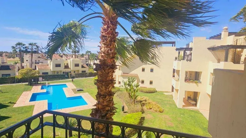 Blick vom Balkon auf die gepflegte Gartenanlage mit großem Pool und Palmen unter blauem Himmel.