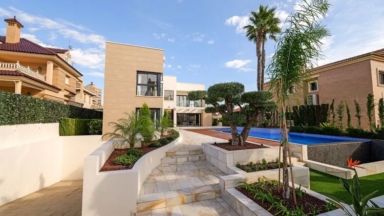 Moderne Villa mit großem Poolbereich, gepflegtem Garten und Palmen unter blauem Himmel.