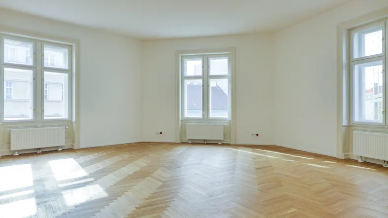 Heller, leerer Raum mit Fischgrätparkett und drei hohen Fenstern, die viel Tageslicht hereinlassen.