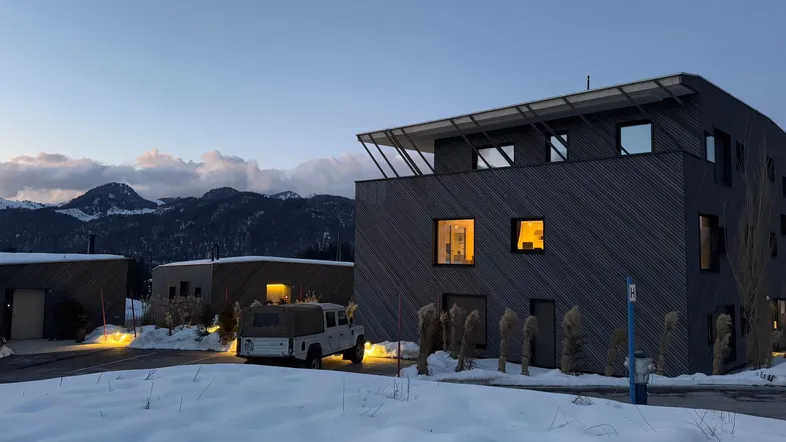 Modernes Gebäude in verschneiter Berglandschaft bei Dämmerung mit beleuchteten Fenstern und einem Geländewagen.