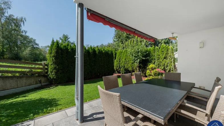 Geräumige Terrasse mit Esstisch und Gartenmöbeln, umgeben von viel Grün und einer Hecke.