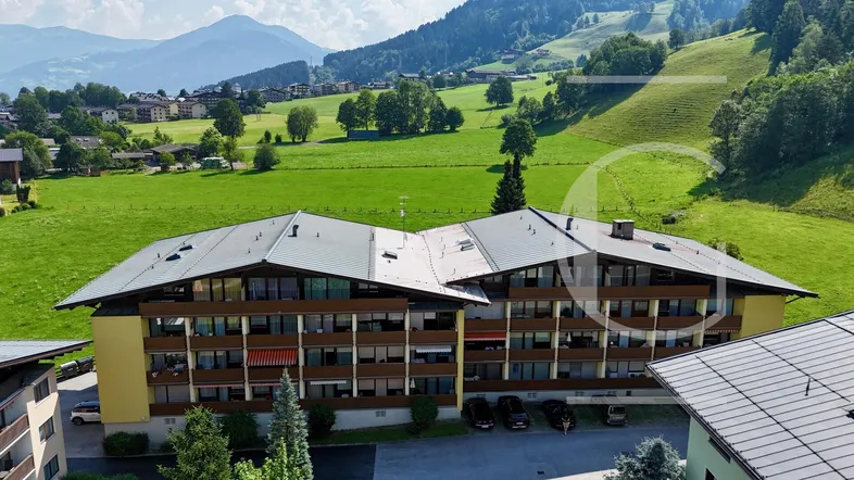 Mehrfamilienhaus in alpiner Landschaft mit Satteldach und Balkonen, umgeben von grünen Hügeln und Bergen.