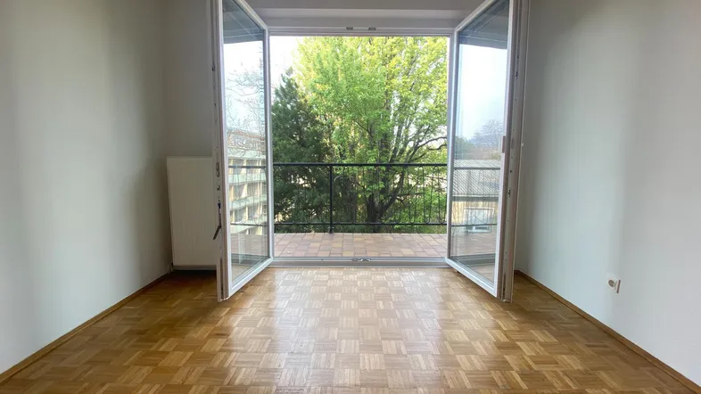 Helles Zimmer mit Parkettboden und Zugang zu einem Balkon mit Blick ins Grüne.