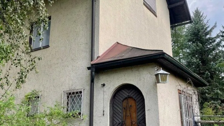 Altes Haus mit Putzfassade, Holztür und Giebeldach, umgeben von Bäumen und Sträuchern.