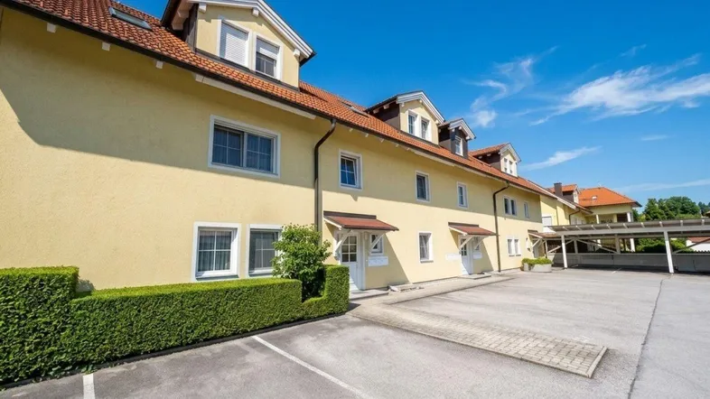 Gelbes Mehrfamilienhaus mit gepflegter Hecke und Parkplätzen unter blauem Himmel.
