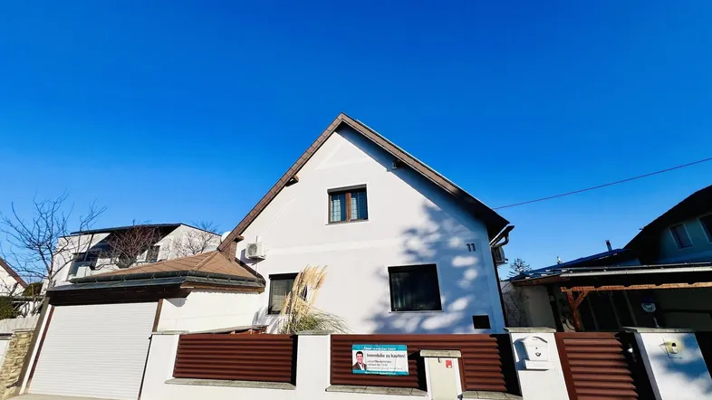 Vorderansicht des Hauses mit weißer Fassade, Satteldach und einem Zaun zur Straße hin.