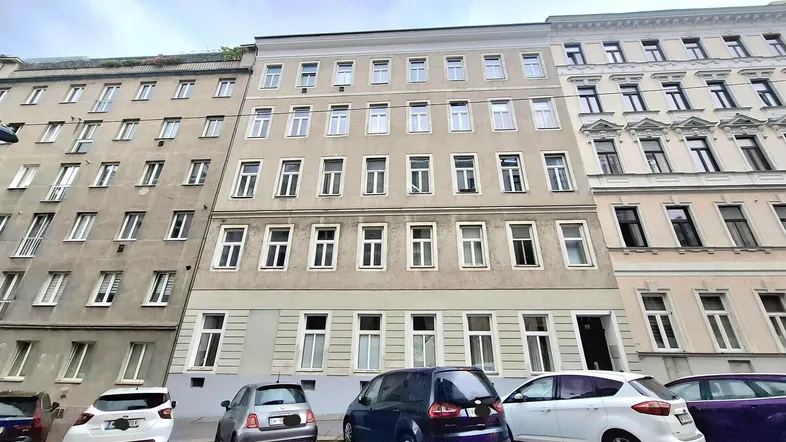 Fassade eines Mehrfamilienhauses in städtischer Umgebung mit parkenden Autos auf der Straße.