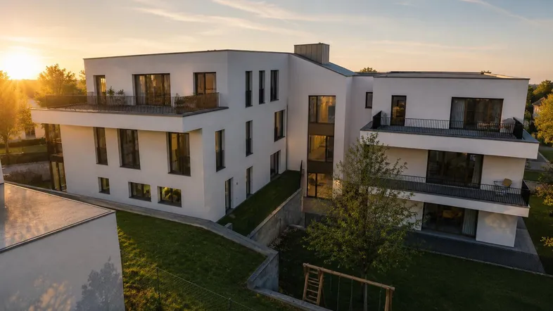 Modernes Mehrfamilienhaus mit Balkonen und Grünflächen bei Sonnenuntergang, umgeben von Natur.