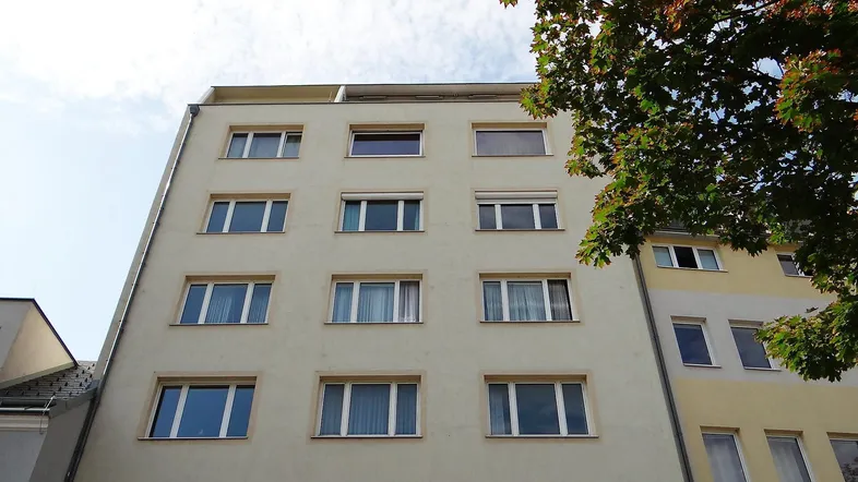 Mehrstöckiges Wohngebäude mit mehreren Fenstern und einem Baum im Vordergrund.