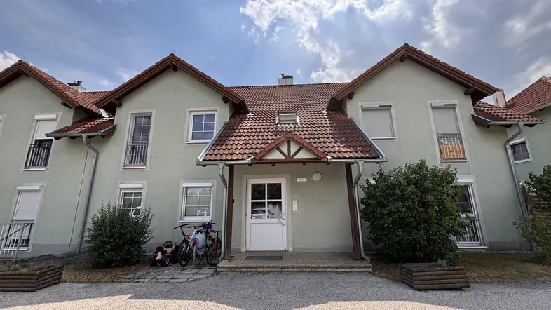 Mehrfamilienhaus mit hellgrüner Fassade, roten Ziegeldächern und kleinen Balkonen unter bewölktem Himmel.