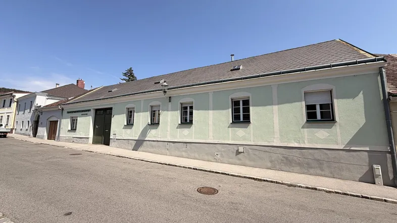 Langes, hellgrünes Gebäude mit Satteldach und mehreren Fenstern an einer Straße gelegen.