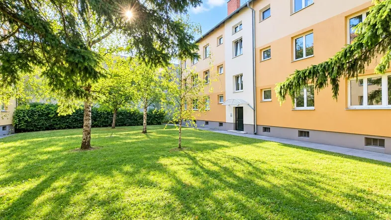 Mehrfamilienhaus mit gepflegter Grünfläche und Bäumen im Vorgarten bei Sonnenschein.