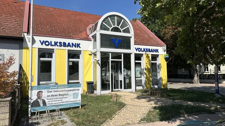 Gelbes Bankgebäude mit rotem Dach und Werbebanner für Immobilienexperten an der Fassade.