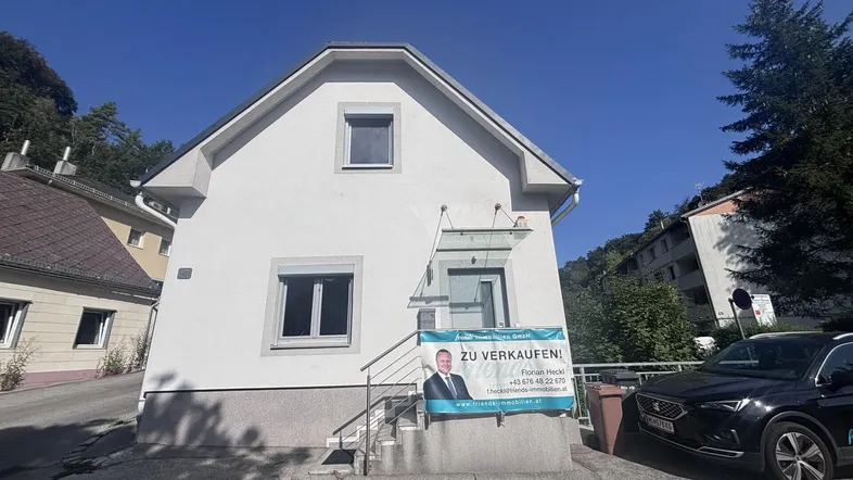 Weißes Einfamilienhaus mit Satteldach und einem "Zu verkaufen"-Schild vor dem Eingang.