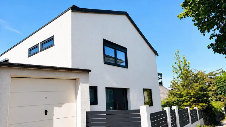 Modernes weißes Einfamilienhaus mit Garage und dunklem Zaun unter blauem Himmel.