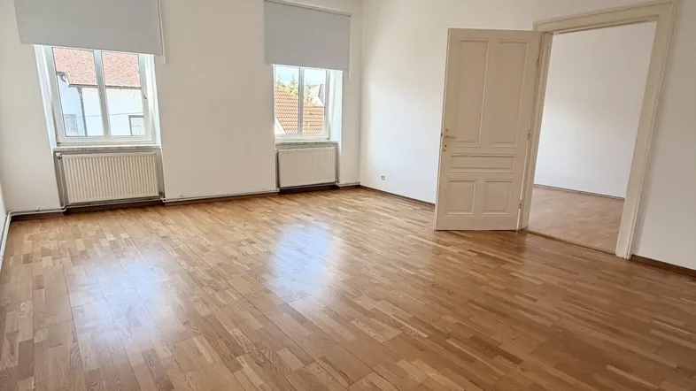 Helles, leeres Zimmer mit Parkettboden, weißen Wänden und zwei Fenstern, bereit zur Einrichtung.