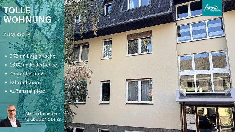 Fassade eines Mehrfamilienhauses mit Fenstern und Eingangsbereich, teilweise von Bäumen verdeckt.