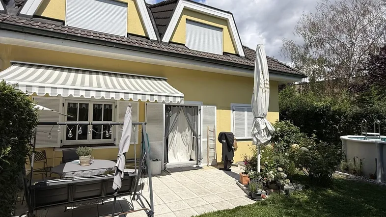 Sonniger Außenbereich mit Terrasse, Markise und Gartenmöbeln vor der gelben Hausfassade.