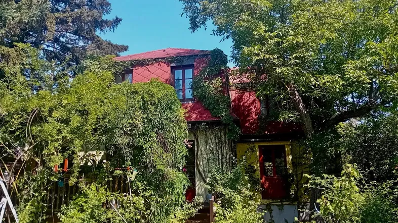 Charmantes Haus mit rotem Dach, umgeben von üppigem Grün und hohen Bäumen.