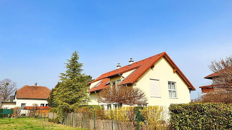 Einfamilienhaus mit gelber Fassade, rotem Ziegeldach und umgebendem Gartenbereich unter blauem Himmel.