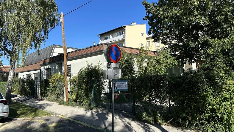Gebäudeansicht von der Straße mit üppiger Begrünung und einem "Zu Kaufen" Schild.
